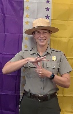 Ranger in uniform stands in front of a hanging purple, white, and gold flag. She signs vote in ASL.
