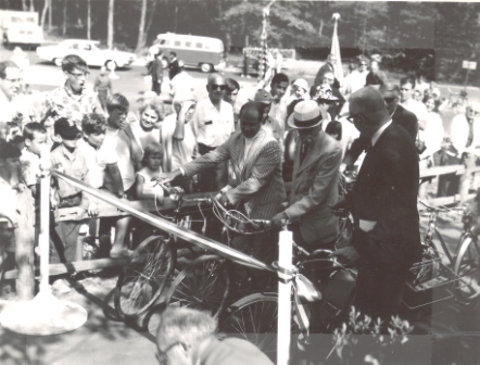 A black and white photo of nicely dressed people from the early 1900s lined up with bicycles behind a ribbon.