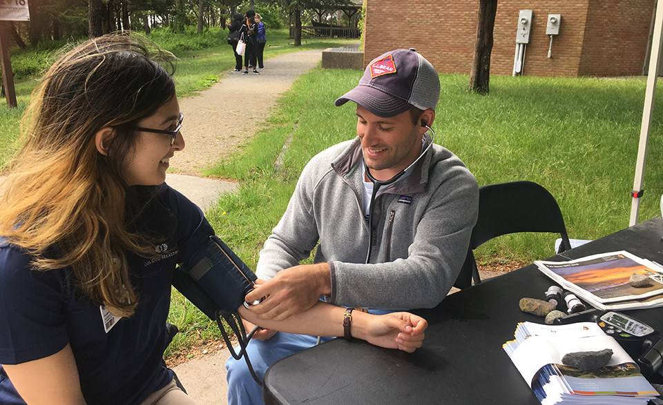 A woman wearing glasses gets her blood pressure checked by a man wearing a ball cap.