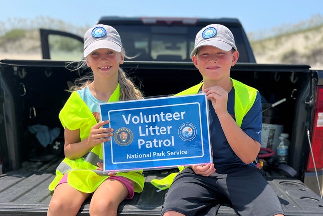 Two children sit on back of black truck dressed in safety vests holding sign that says Volunteer Litter Patrol