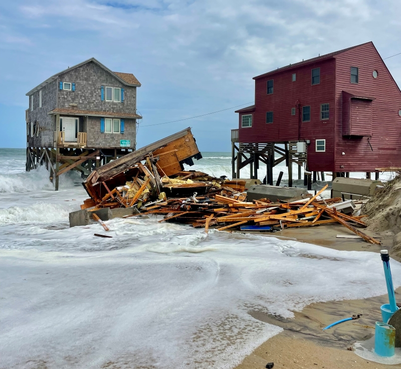 Debris on the beach and in the water in front of a house on pilings in the ocean.