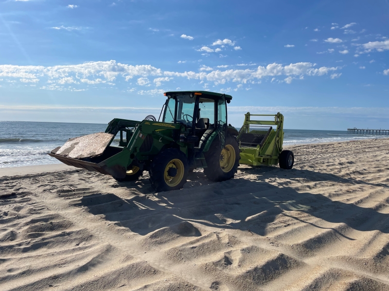 Photo of a green tractor and surf rake on the beach on a sunny day.