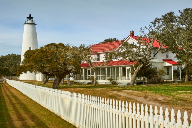 White fence with white lighthouse and white house with red metal roof.