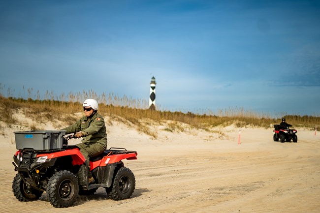 Two ATV riders drive on the beach. The Cape Lookout Lighthouse is in the background, between the two riders.