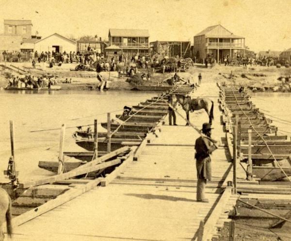Black soldier stands on pontoon bridge over river with buildings and groups of people in background