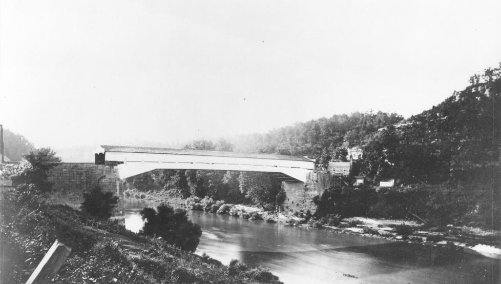 A black-and-white photo of a white, wooden covered bridge over a river.