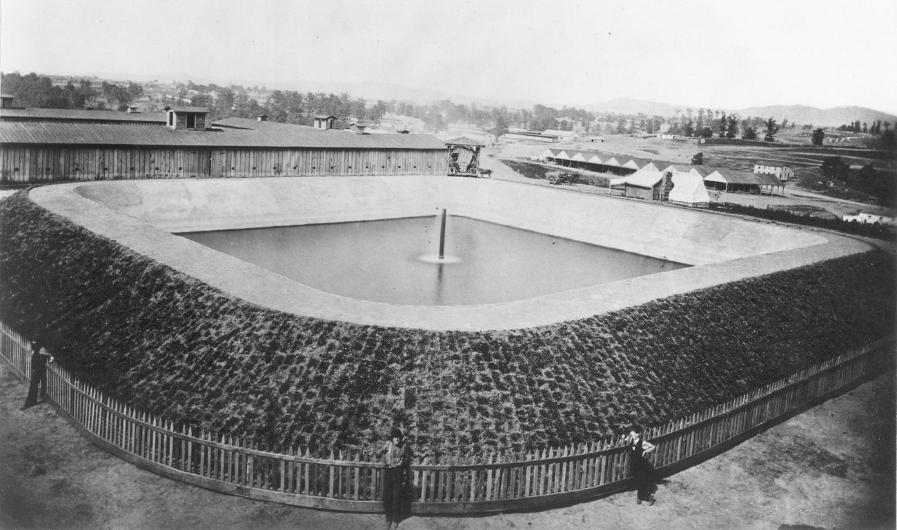 Civil War earthen reservoir that held reservoir with fountain. A wooden fence encloses the reservoir. Wooden buildings can be seen in the background.