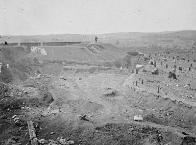 A large Civil War earthen fort with a soldier standing on the top parapet, to the right in front of the fort is a dry ditch and tree stumps. Hill cans been seen in the background.