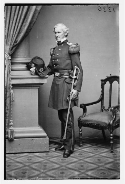 Portrait of Brigadier General Lorenzo Thomas in uniform during the Civil War. He is standing in a studio next to a chair with a hat in his right hand and sword in his left.