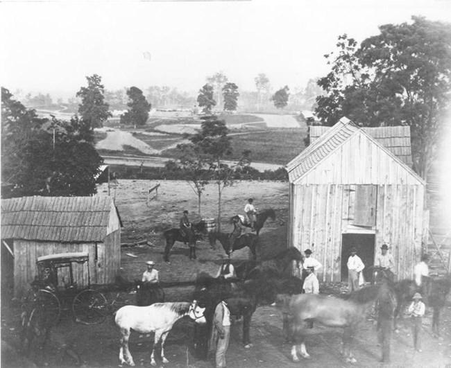 Men and horses in front near wooden buildings with earthen fortification in the background during the Civil War.