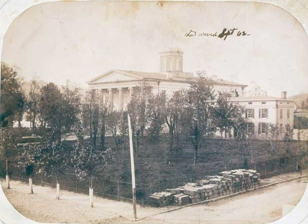 Large state house building in background with field, trees, and fence in front during the Civil War.