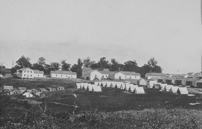 Tents and wooden hunts in foreground with many wooden cottages and larger buildings in background during the Civil War.