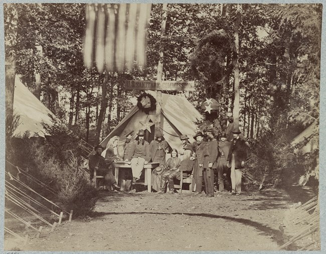 A group of soldiers and civilian standing and sitting in front of a tent with a sign that reads "U.S. Sanitary Commission."