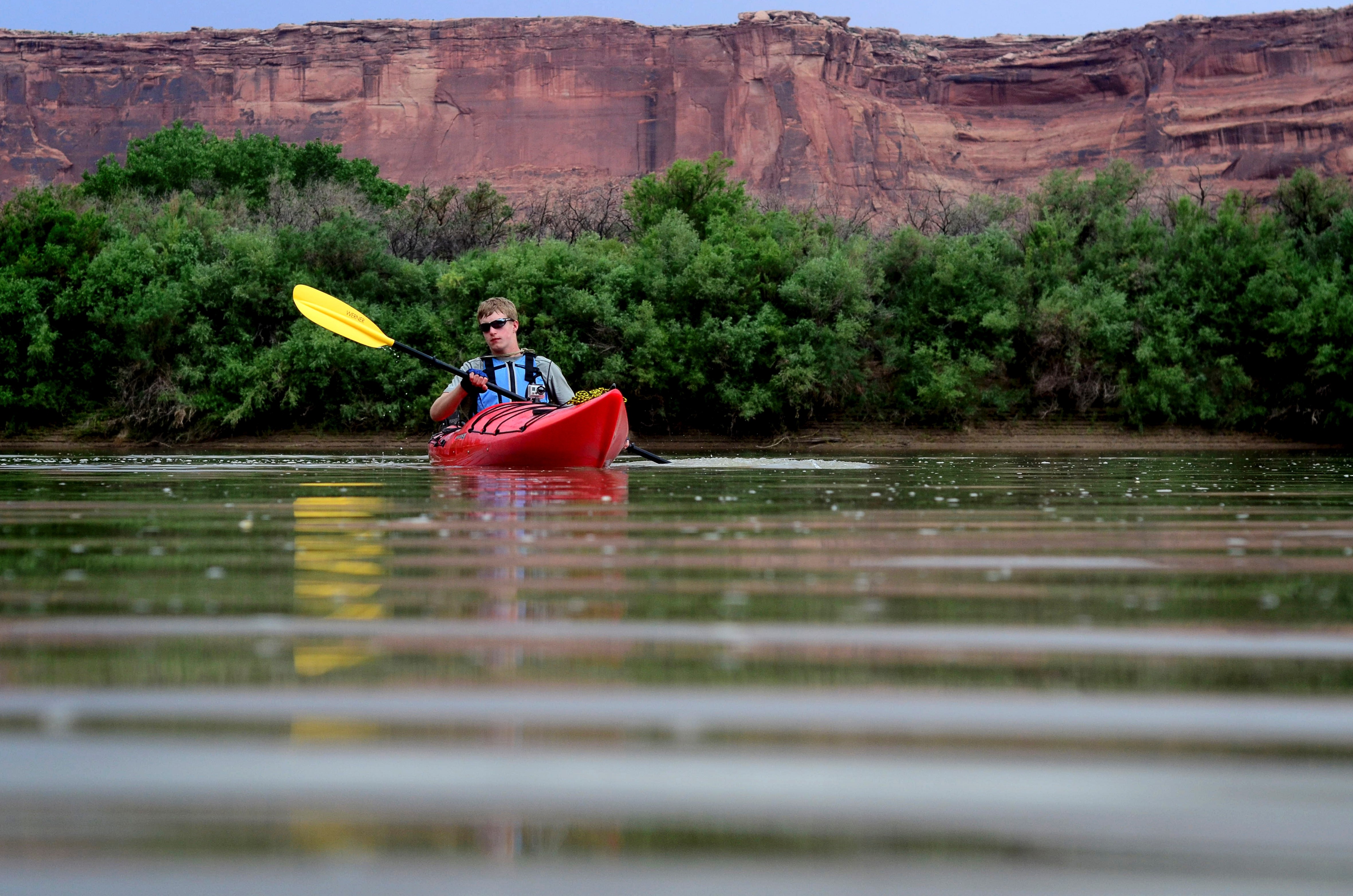 light-skinned man in red kayak paddles on a river, red canyon wall and green shrubs in background