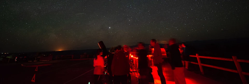 people illuminated with red lights stand near a telescope with a starry sky overhead