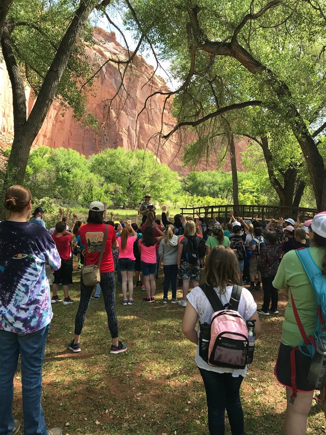 Large group of children and adults face a park ranger standing under red cliffs, large green trees, and blue sky.