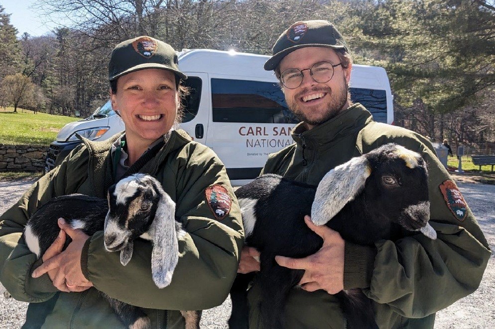 Two park rangers hold two baby goats in their arms