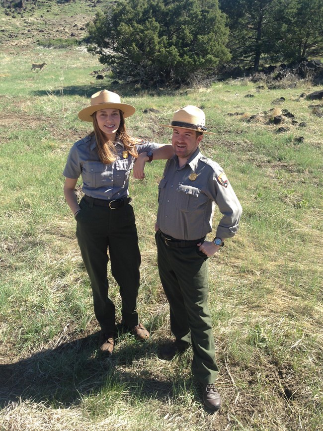 A female and male park ranger stand in a grassy area on a sunny day.  The female ranger on left rests her elbow on the male ranger's shoulder to the right.  They both smile facing the camera.  A deer in the right background makes its way toward the trees.