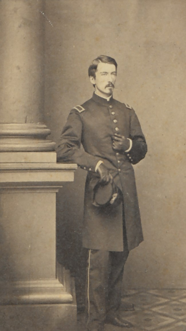 A young 1860s army officer stands next to a pillar for a full portrait photograph.