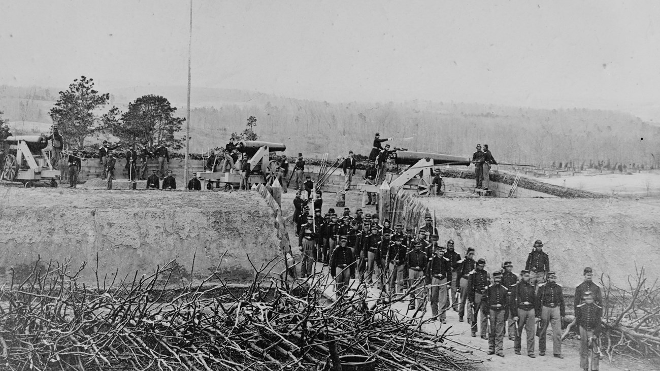 A column of soldiers marches forth from an earthwork fort with cannons.