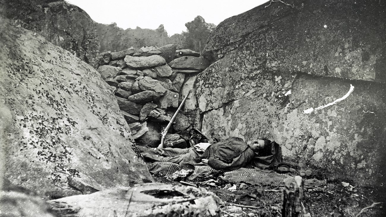 An 1863 photo in stark black and white depicts a dead US soldier in a trench fortified by rocks.