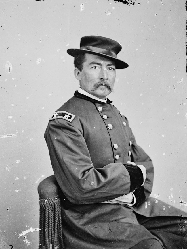 An 1864 portrait photo shows a mustached man seated, hand-in-waistcoat, in an army uniform.