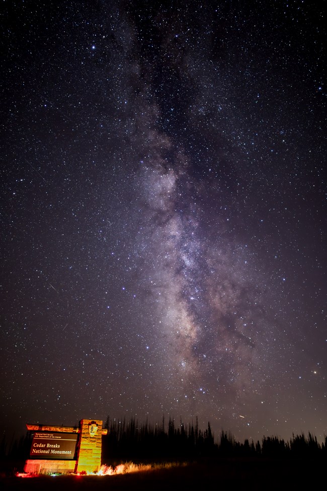 The Cedar Breaks National Monument entry sign illuminated at night beneath a sky filled with stars.
