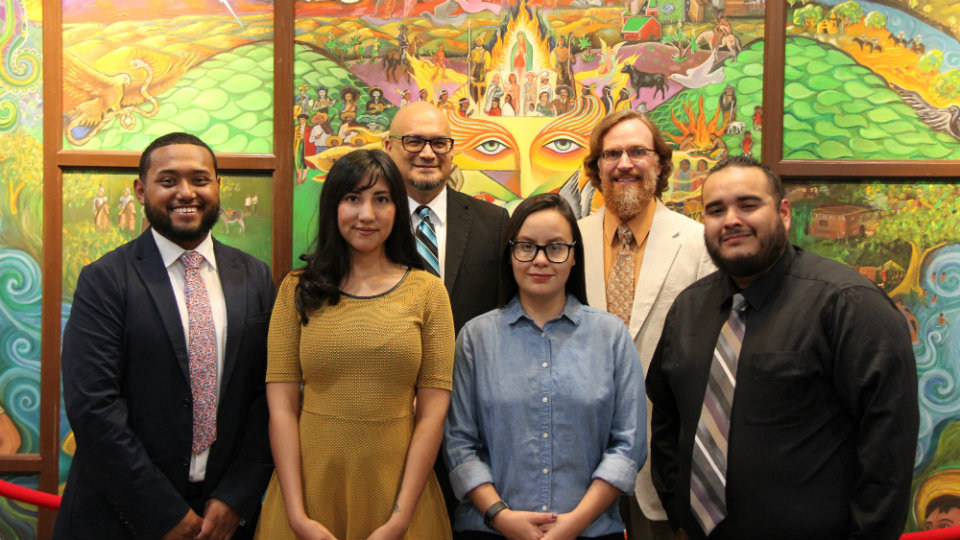 Four men, two women standing in front of brightly-colored mural facing you