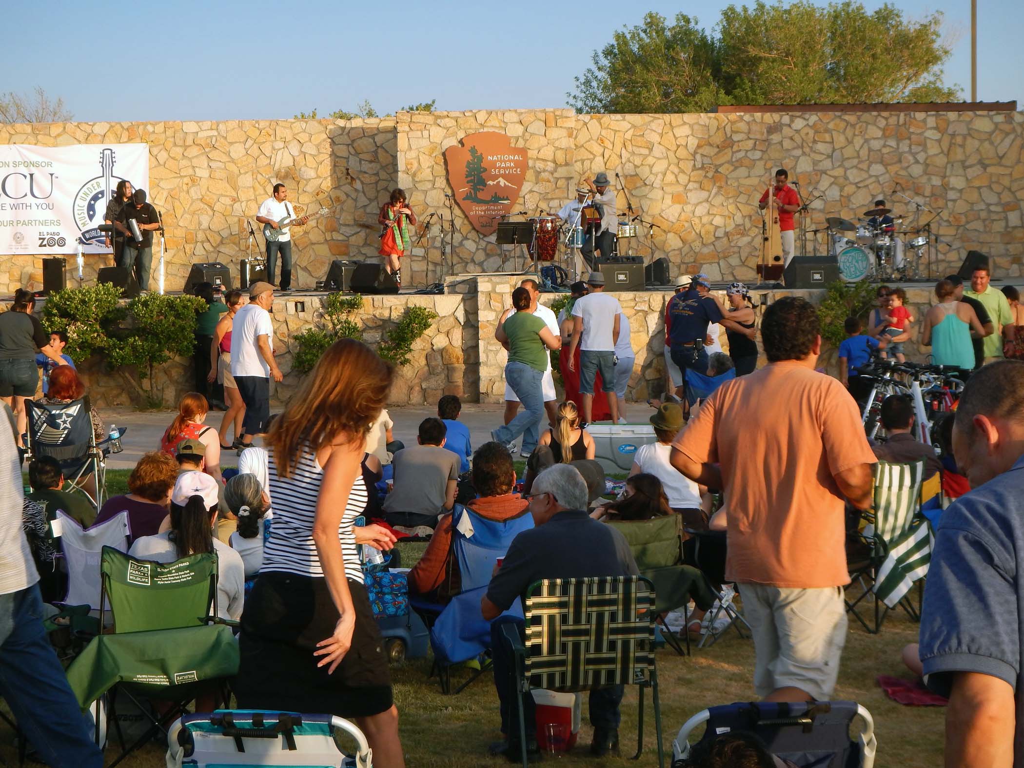 the back of a large group of people, some sitting in lawn chairs and some dancing, in front of amphitheater with band onstage