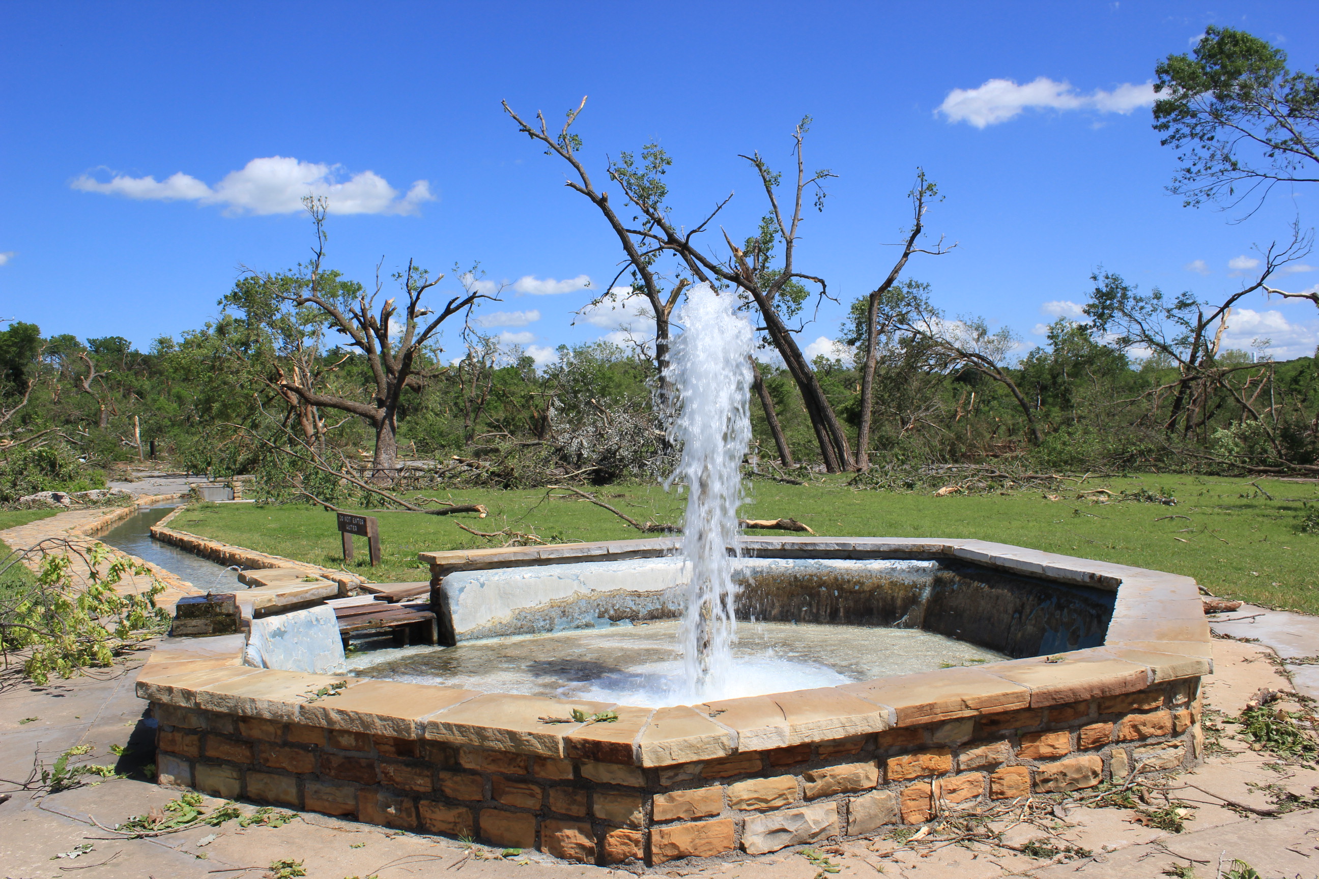 A jet of water rises out of a stone circular basin. Broken trees and limbs are in the area behind and around the basin