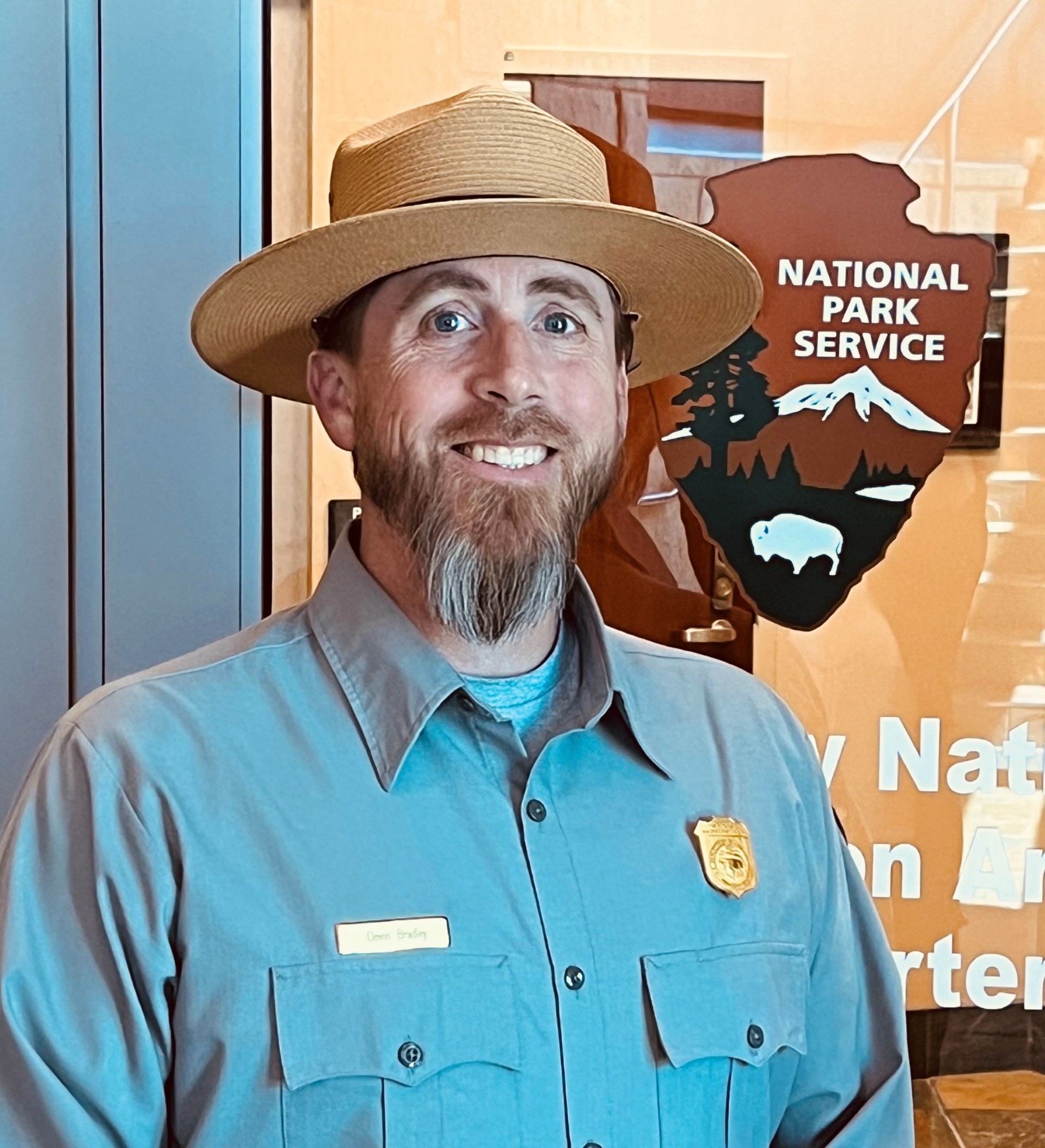 a smiling man in a ranger hat and a grey uniform shirt stands near a park service arrowhead logo