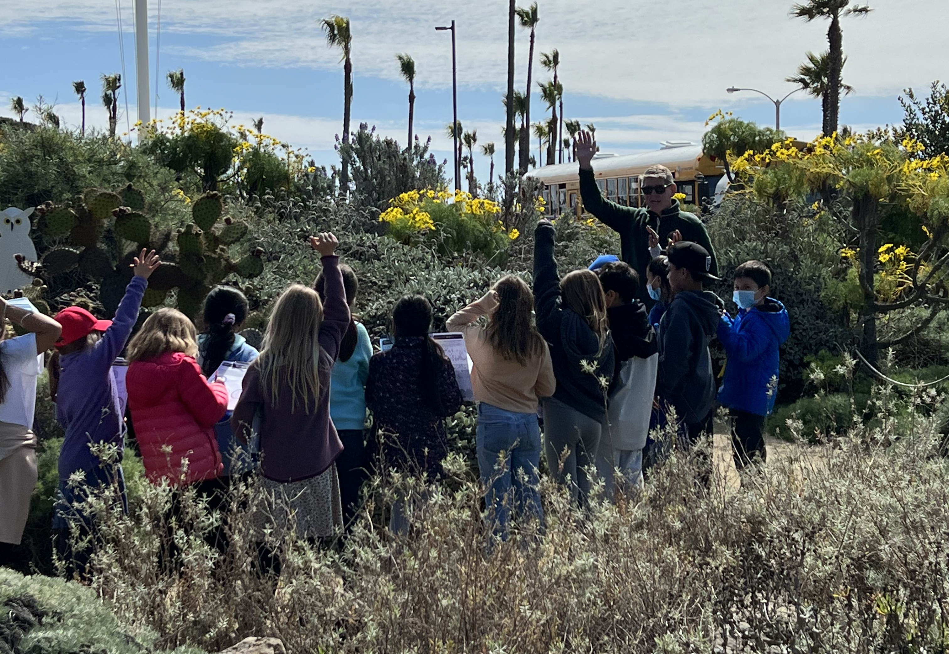 A group a students explore a garden with a male volunteer ranger. A school bus and American flag are in the background.