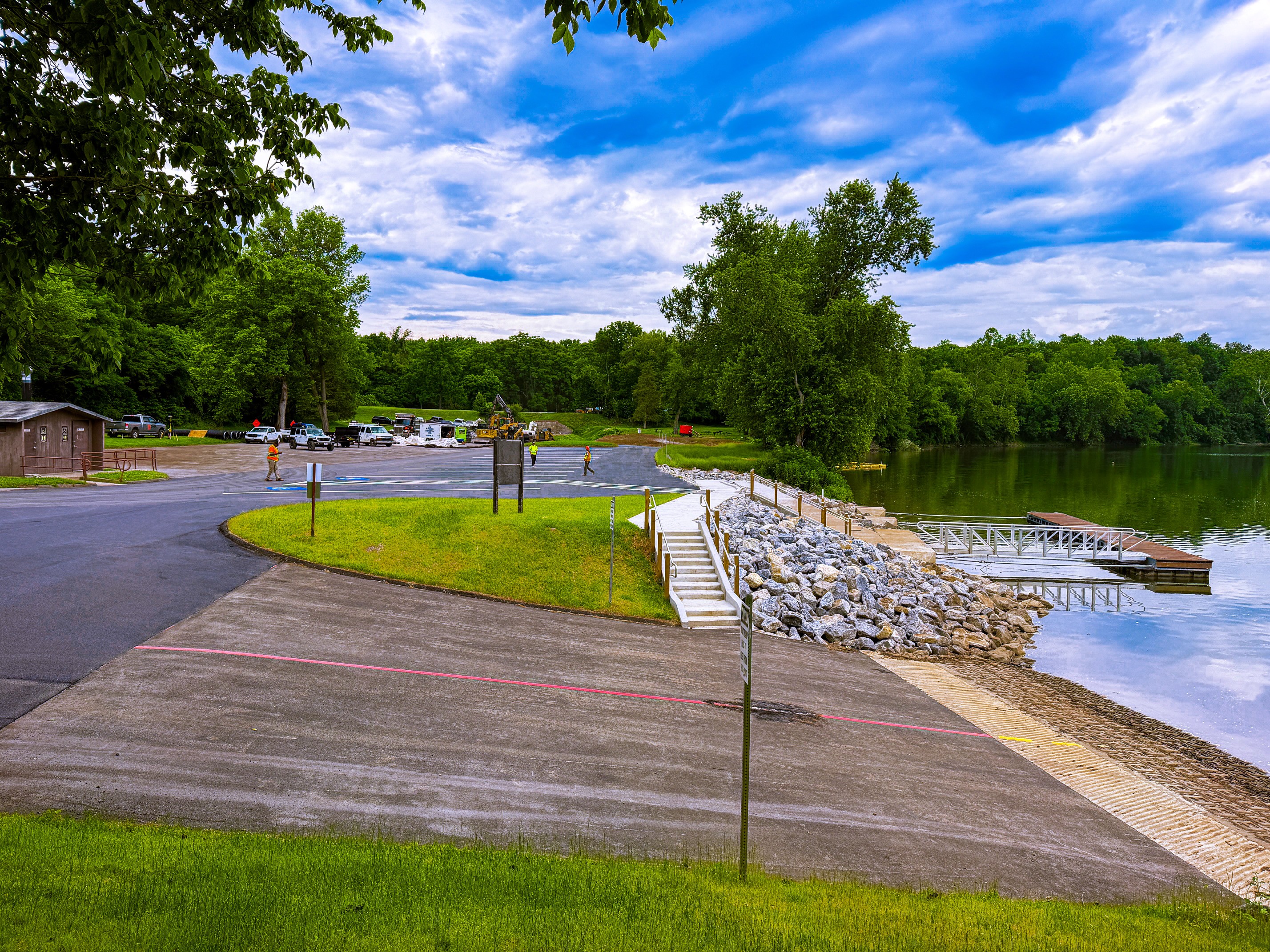 A concrete boat ramp extends down to a river, with a blue sky and green trees in the background