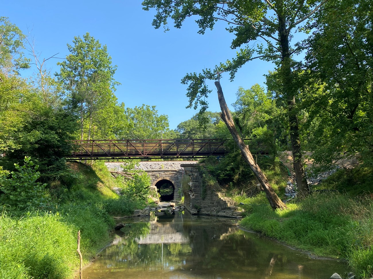 A brown metal bridge spans the towpath trail across the remnants of a masonry culvert