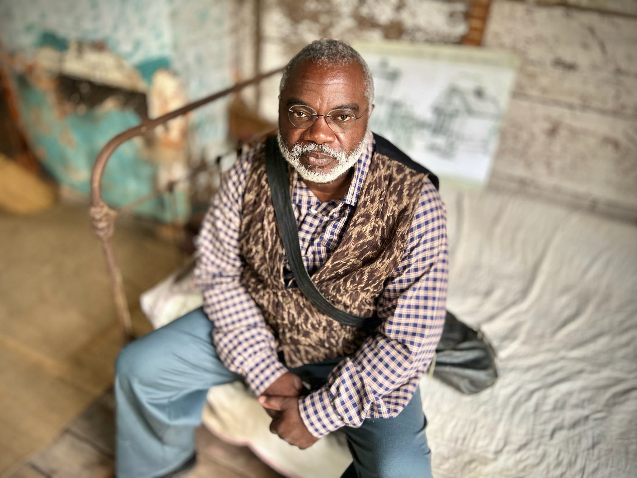 An African American man wearing glasses is seated on a bed in a room; he wears glasses and has a bag slung across his shoulder. He is dressed in a brown vest over a plaid long-sleeve shirt, and blue pants.