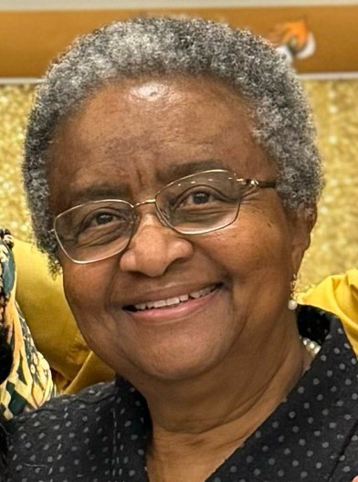 A woman is shown with curly grey hair and glasses while smiling.