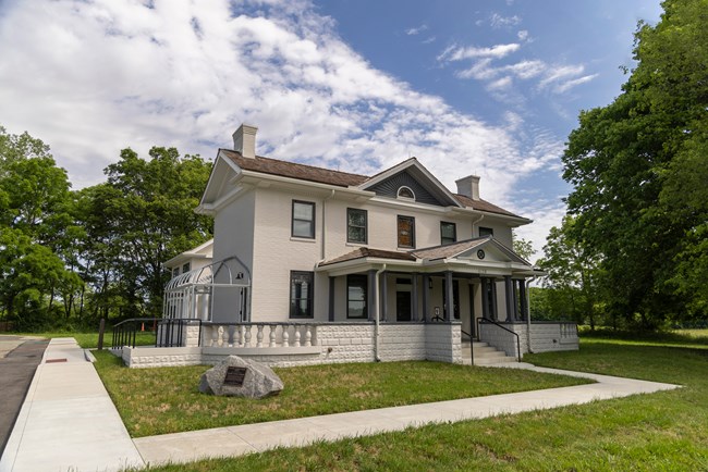 light gray two story brick building with a porch that is covered by a roof with dark gray pillars holding up the roof. There is green grass in the foreground. A cement sidewalk goes to the front of the porch stairs that leads into the home.