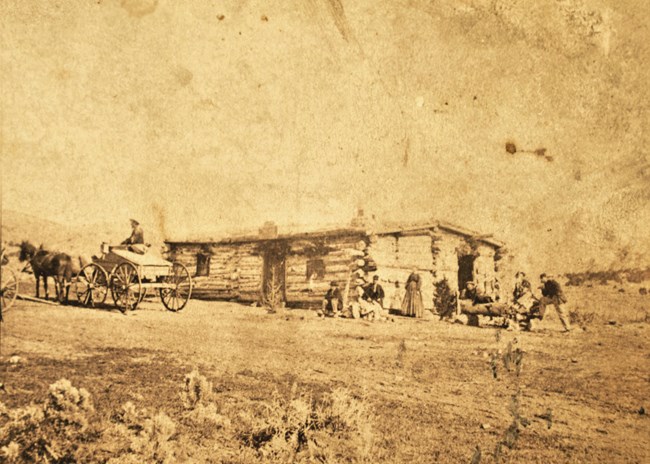 Historical Sepia photograph depicting a log building with several people sitting out front with a man sitting in a single horse drawn wagon.