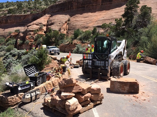 Equipment and pallets of large sandstone rock in the foreground await shaping and placement. In the background 6 staff members are working to install new rock to form walls along road.