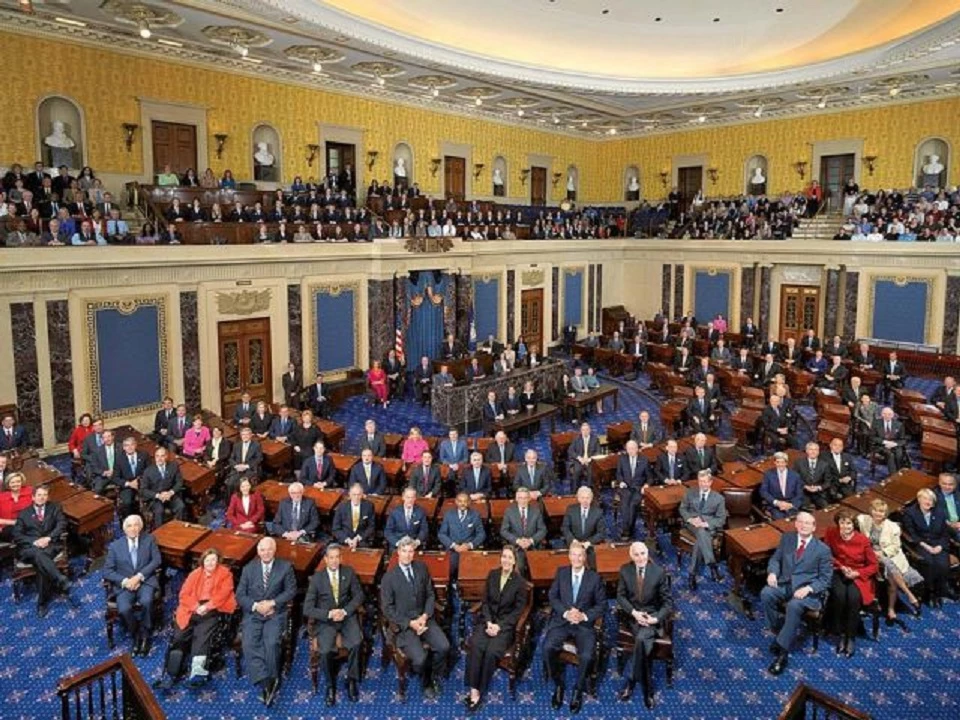 Senators seated in the Senate chamber. Dark wood desks form a semi-circle around the room