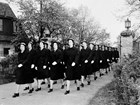 A group of uniformed women march down the road between a house and a lampost