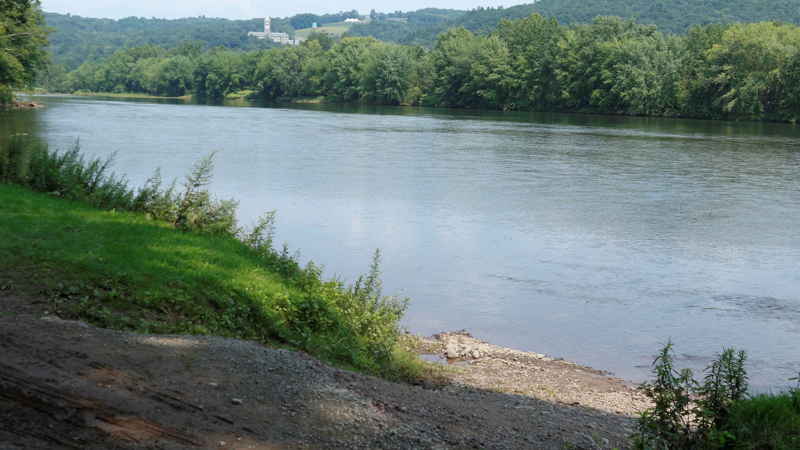 River access with gravel ramp into river. Lush trees line bank. Tree-covered hills in distance.