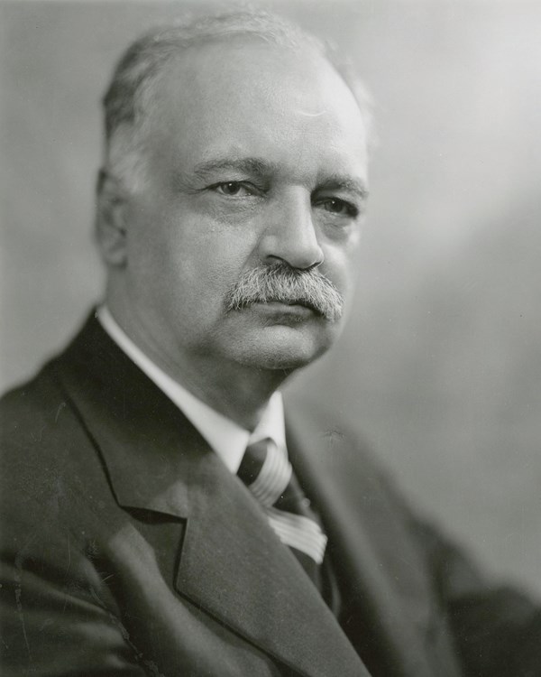 A 1928 bust portrait photo shows a serious gray haired and mustached man in a suit.