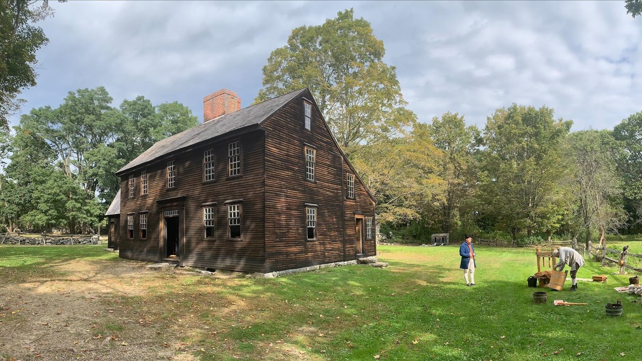 A wooden house sits in an open grassy field. Three people in colonial clothing press cider in front.