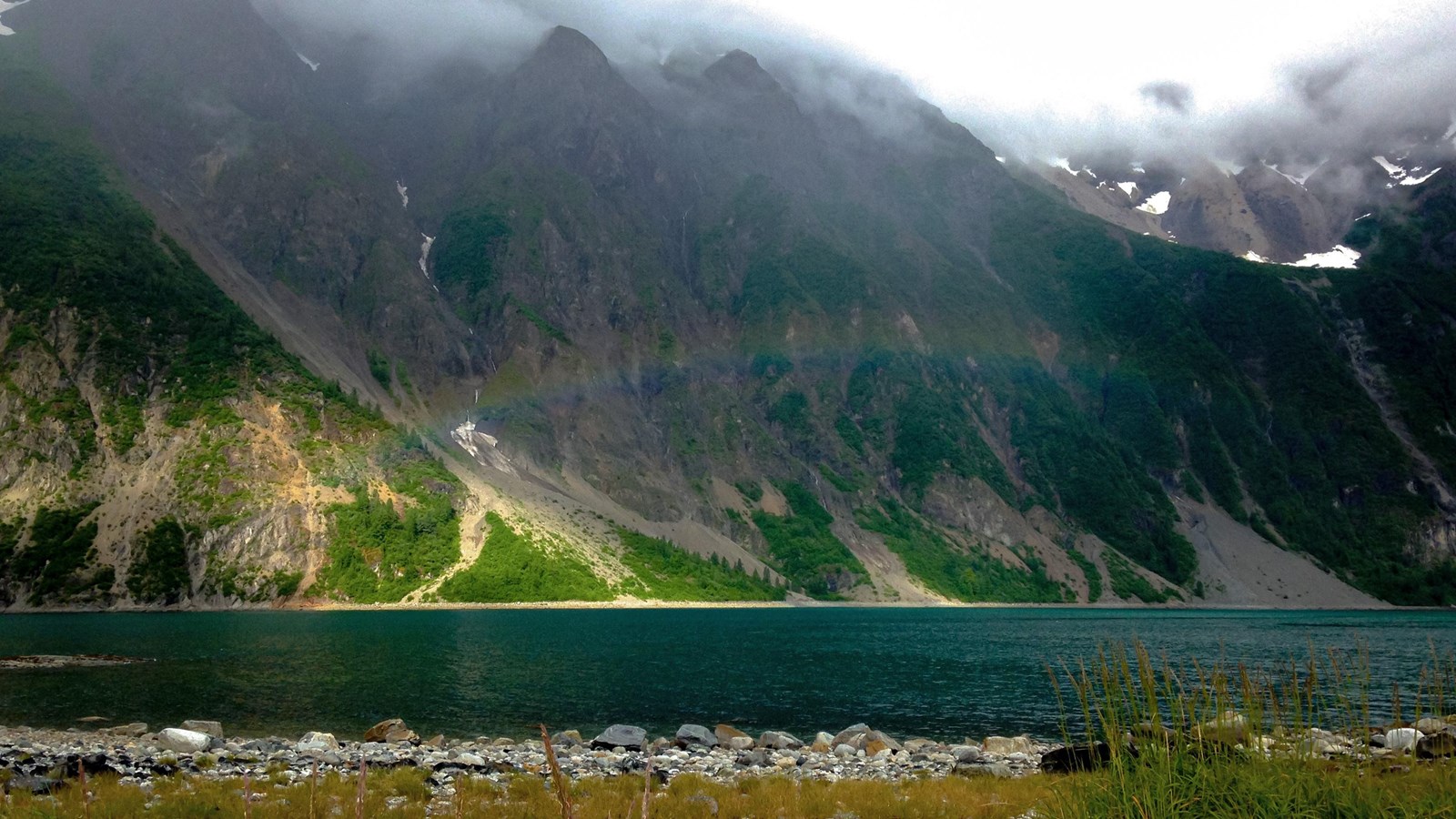 a rainbow stretches above water before a steep mountainous cliff on a cloudy day.