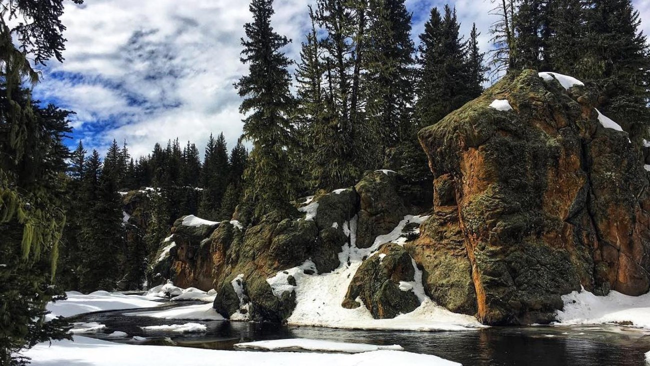 A snow-covered landscape with a small stream meandering through rock spires and evergreen trees.