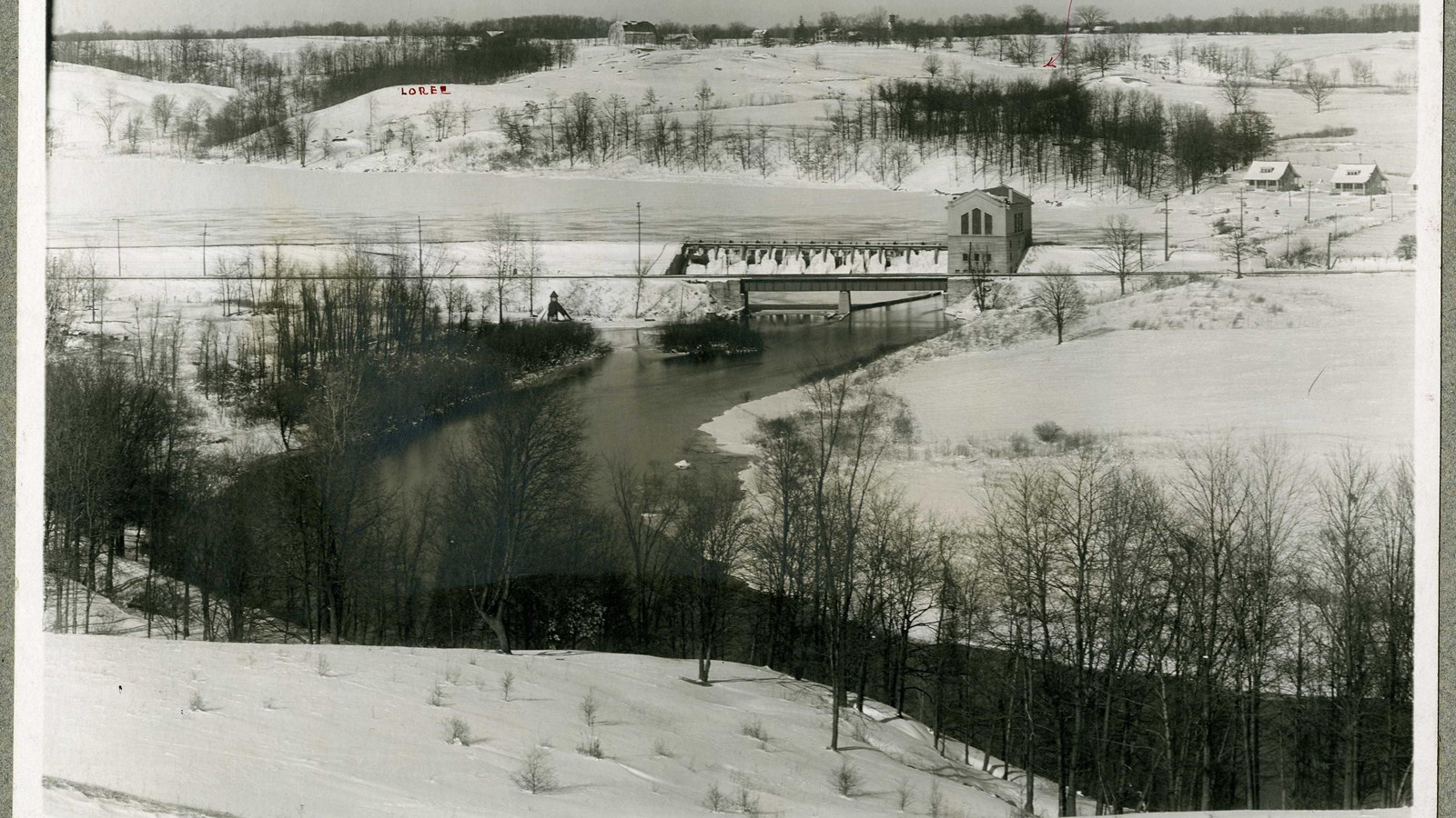 Black and white of hilly area covered in snow with river and bridge and building in distance.