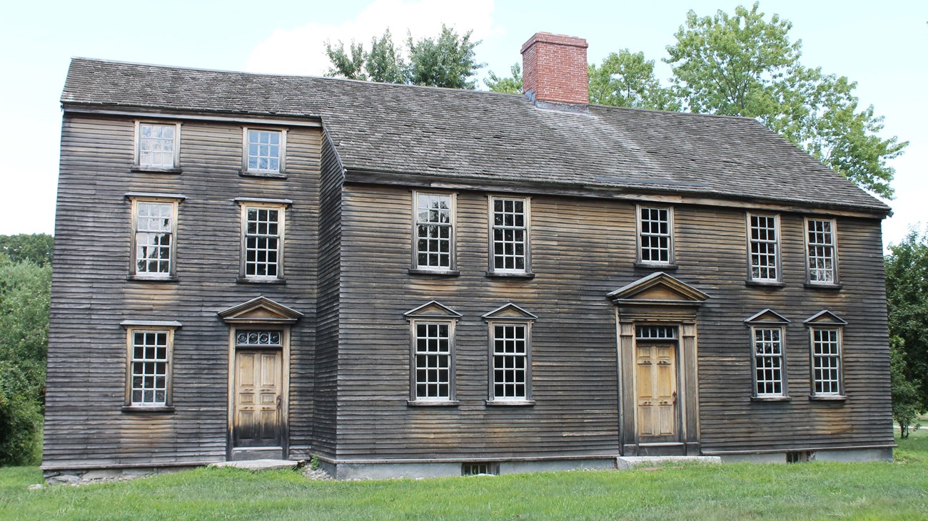 A wooden 2.5 story house with central chimney and 8/8 pane glass windows has a green lawn in front.