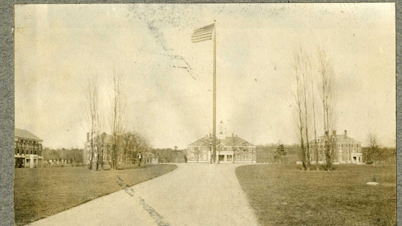 Black and white of road leading to group of buildings with flag pole in middle, trees on edge