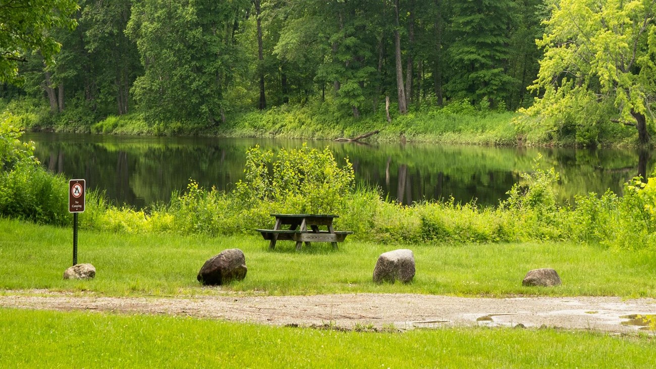 Available picnic tables sit on a grassy area next to a river.